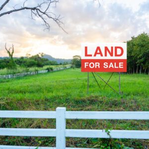 A land for sale sign on empty land surrounded by a white fence and trees.